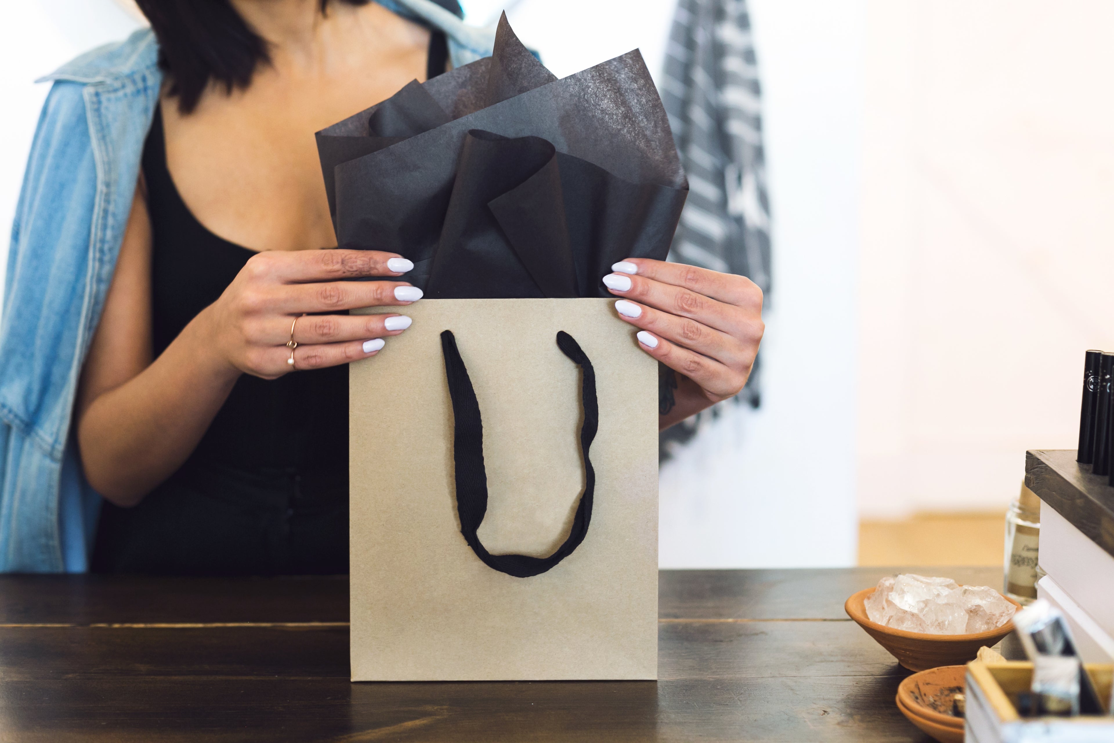 A woman with white nails holding a Natural tan square shopping bag with black handles and black tissue paper