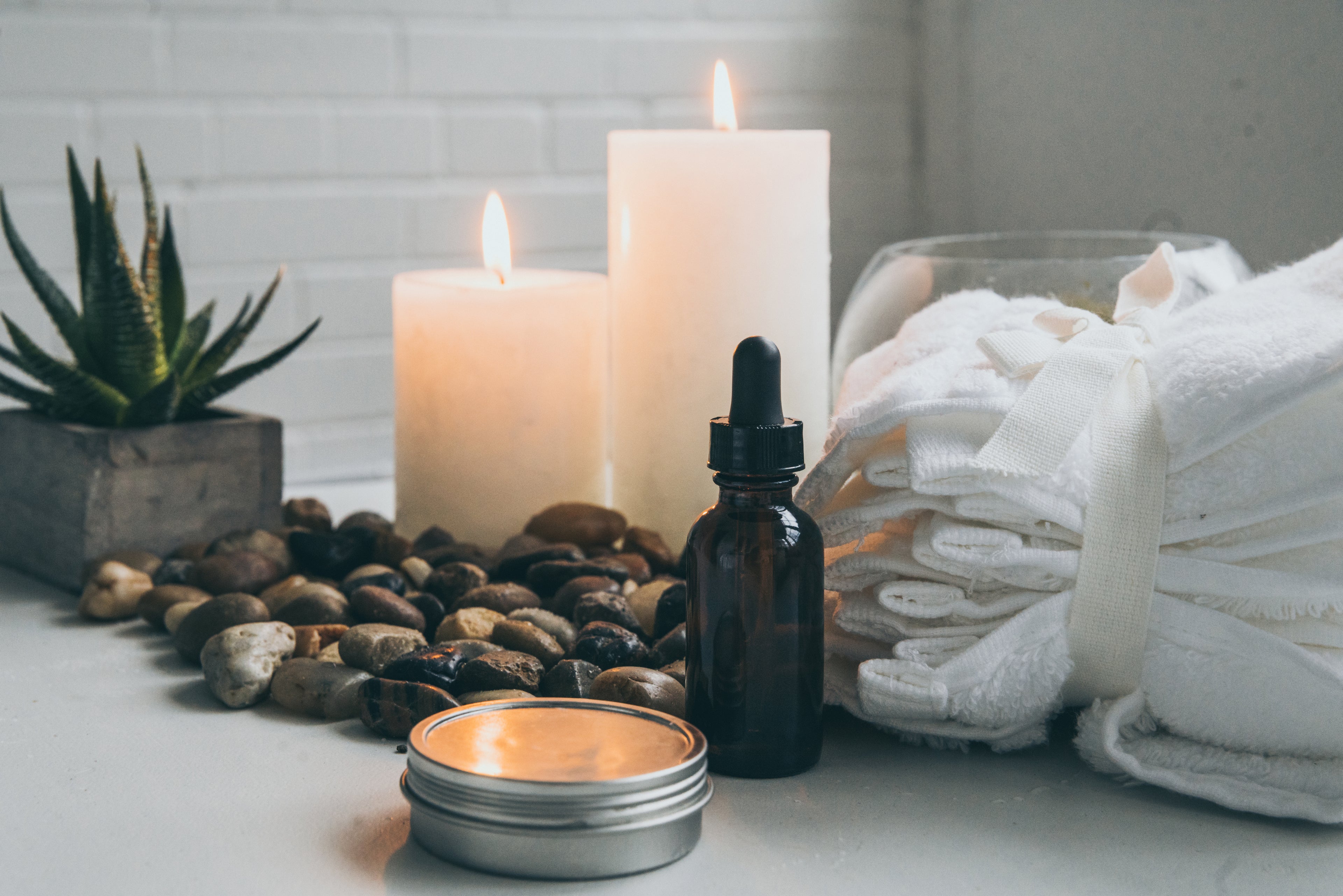 Natural supplies for the spa including a candle, a metal tin, rocks, aloe plant, stack of white towels and an amber colored glass dropper bottle.